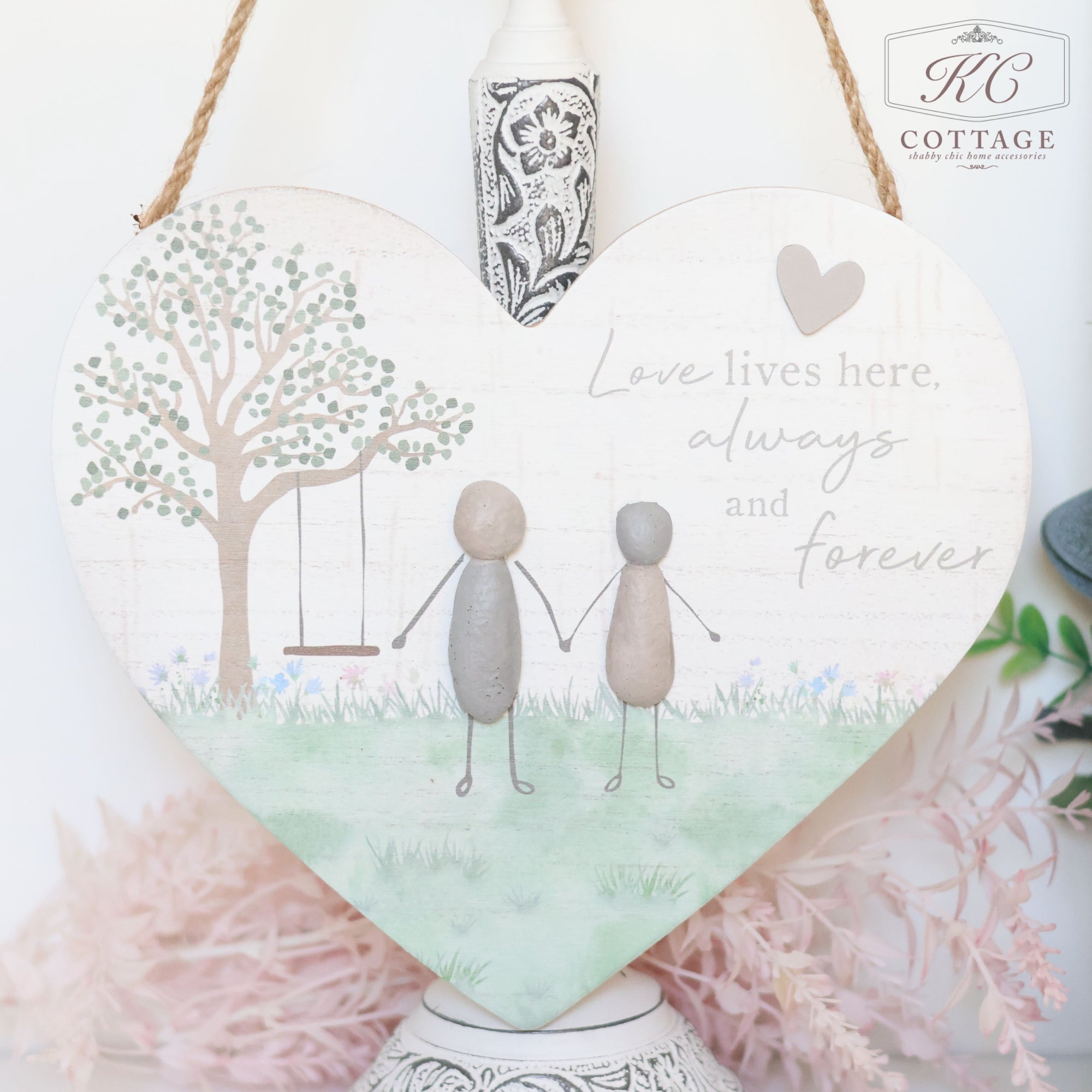 A Pebble Friends & Love Heart Plaque with a painted scene of two pebble figures holding hands under a tree with a swing. The background features soft pastel greenery and flowers. Sentimental quotes read, "Love lives here, always and forever." This charming piece of home decor is hung by a rope.