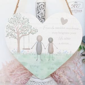 A heart-shaped decorative sign featuring a tree with a swing and two characters made of pebbles holding hands. The sign reads, "Friends are like rainbows, they brighten your life after a storm." The background has artistic flower and grass elements, with a delicate pink floral arrangement below. Pebble Friends & Love Heart Plaques are perfect for adding sentimental quotes to your home decor.