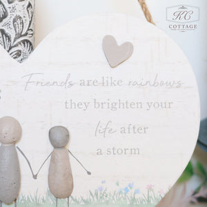 A heart-shaped wooden plaque with an illustration of two stick figures holding hands. The text on the plaque reads, "Friends are like rainbows, they brighten your life after a storm." A small heart is above the text, and the bottom edge is adorned with tiny flowers. Perfect for home decor and adding sentimental quotes to your space, this can be found under Pebble Friends & Love Heart Plaques.