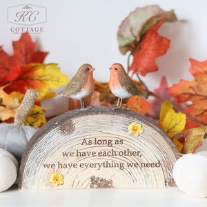 The Autumn Robin Couple On Resin Bark Base features two small bird figurines perched on a log-shaped block inscribed with the sentiment, "As long as we have each other, we have everything we need." The backdrop boasts vibrant red, orange, and yellow autumn leaves along with charming white pumpkins—a whimsical piece ideal for nature enthusiasts.