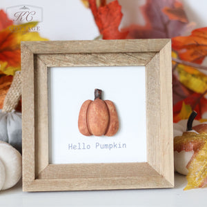 The Autumn Pumpkin 3D Framed Plaque With Quotes features a simple illustration of an orange pumpkin with the text "Hello Pumpkin" underneath. The small wooden frame is surrounded by autumn leaves in orange and red hues, along with small white pumpkins. Perfect for Pumpkin Decor, the "KC Cottage" logo graces the upper left corner.