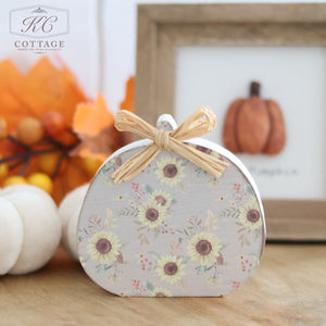 A charming Autumn Rustic Design Wooden Pumpkin with a raffia bow sits on a wooden surface. Behind it, vibrant orange leaves and a framed pumpkin picture enhance the cozy fall decor. The scene radiates an autumn rustic design, while a logo at the top reads "KC Cottage.