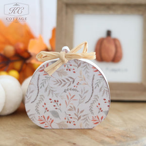 A charming, pumpkin-shaped box featuring an intricate floral design and adorned with a raffia bow rests on a wooden surface. In the background, a framed picture showcasing the "Autumn Rustic Design Wooden Pumpkins With Raffia Bow" is surrounded by fall decorations, including orange leaves and small pumpkins, enhancing the rustic autumn ambiance.