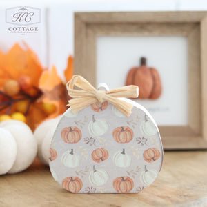 The Autumn Rustic Design Wooden Pumpkins With Raffia Bow perfectly embodies a small decorative item shaped like a pumpkin, covered in a pattern of orange and white pumpkins with green leaves. It is topped with a charming raffia bow. Behind it sits another pumpkin decoration inside a wooden frame, surrounded by fall decor with autumn leaves and white pumpkins in the background.