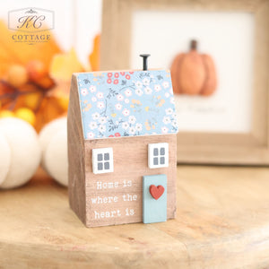 The Wooden House With Floral Roof is a quaint decorative piece featuring a floral-patterned blue roof embellished with flowers and a red heart on its blue door. It is inscribed with "Home is where the heart is" and makes a charming addition to your autumn décor, nestled among other seasonal items like a framed picture of a pumpkin.