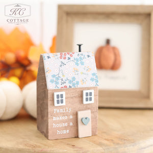 A miniature wooden house with a floral roof, titled "Wooden House With Floral Roof," bears the phrase "Family makes a house a home." Positioned behind this charming ornament is a framed picture showcasing a pumpkin, surrounded by autumn leaves that enhance the cozy, fall-themed decor.