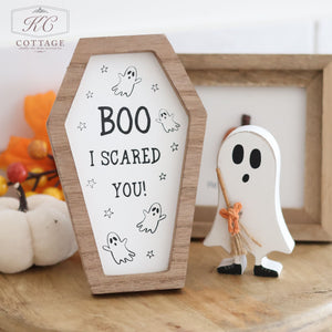 The Halloween Coffin Shaped Signs, crafted from high-quality wood and featuring the text "BOO I SCARED YOU!" along with ghost illustrations, are placed on a table. Beside them stands a small wooden ghost decoration holding a broom, surrounded by decorative pumpkins and autumn leaves. This spooky Halloween decor brings charm to any space.