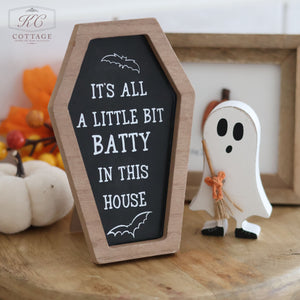 A spooky Halloween decor piece, the Halloween Coffin Shaped Sign, features a high-quality wood sign shaped like a coffin. It is inscribed with "IT'S ALL A LITTLE BIT BATTY IN THIS HOUSE" and flanked by two bat illustrations. Accompanying the sign is a charming ghost figurine clutching a straw broom.