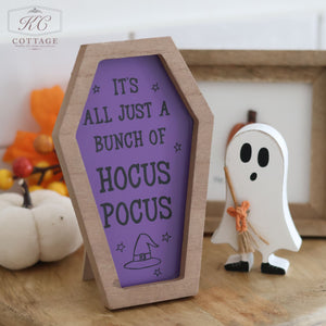 A spooky Halloween decor display features a high-quality wood, coffin-shaped sign from the Halloween Coffin Shaped Signs collection, with the text "IT’S ALL JUST A BUNCH OF HOCUS POCUS" written on it in purple. Next to the sign is a small wooden ghost decoration holding a broom, complemented by autumnal decorations in the background.