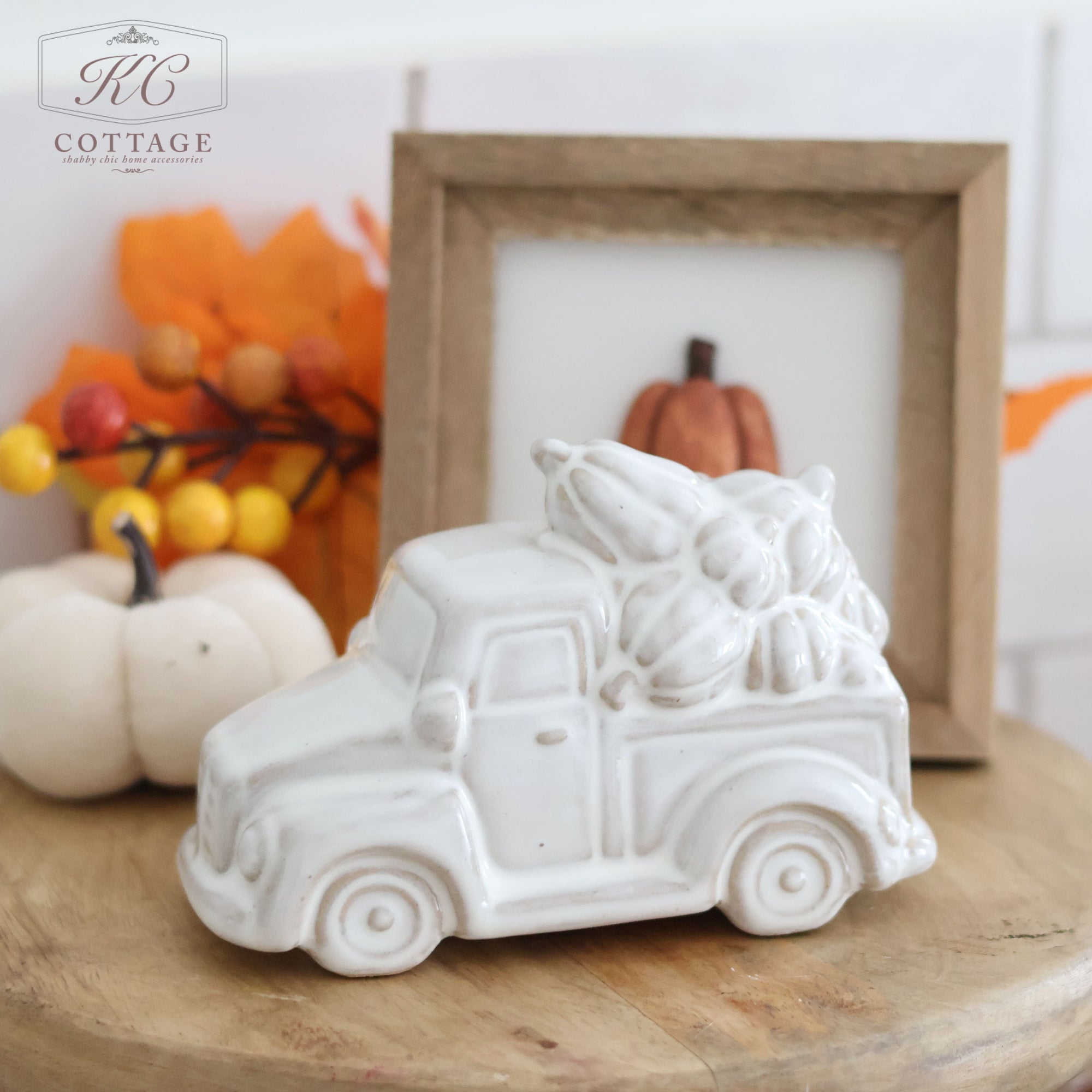 A white **Ceramic Pumpkin Truck Ornament** carrying pumpkins in its bed is perched on a wooden surface. Behind it, a small framed picture of a pumpkin and various fall decor elements—including white pumpkins and orange leaves—create an inviting autumnal scene, ideal for cozy seasonal home decor.