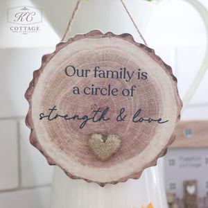 The "Family Bark Hanger With Resin Pebble Heart" is a wooden slice plaque hanging from a string, inscribed with the phrase "Our family is a circle of strength & love," and adorned with a resin pebble heart decoration below the text. This charming piece, ideal for cozy home decor, features the KC Cottage logo in the top left corner.