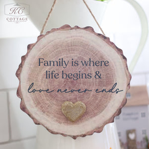 The Family Bark Hanger With Resin Pebble Heart is a charming wooden decoration in the shape of a tree slice. It features a glittery resin pebble heart at the bottom center and bears the heartfelt message, "Family is where life begins & love never ends." Hanging against a white background with the KC Cottage logo visible in the top left corner, it’s perfect for adding warmth to your home decor.