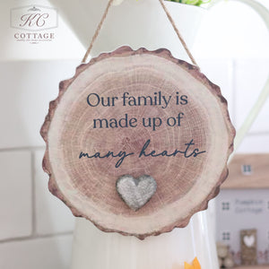 The Family Bark Hanger With Resin Pebble Heart, featuring the text "Our family is made up of many hearts," hangs gracefully with a resin pebble heart below. Displayed in a light, airy kitchen setting adorned with small decorative elements, it brings warmth and love to your home decor.