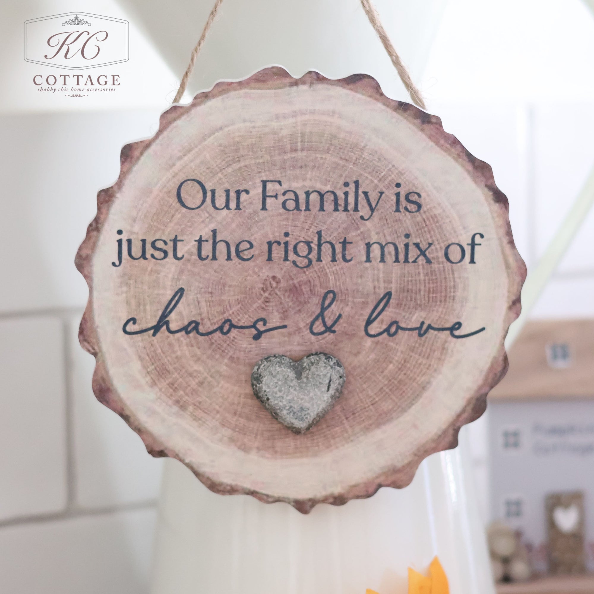 The Family Bark Hanger With Resin Pebble Heart, a decorative wooden sign shaped like a tree slice and perfect for home decor, hangs from a string. It features the text "Our Family is just the right mix of chaos & love" in black font with a resin pebble heart below, and is adorned with the "KC Cottage" logo in the top left corner.