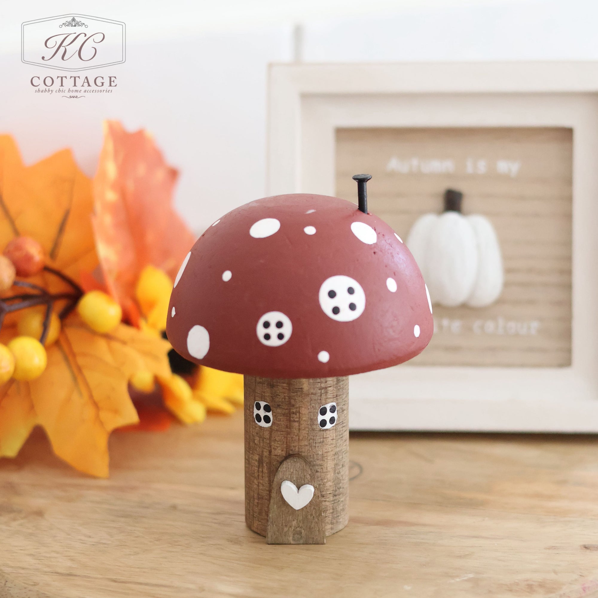 The Autumn Dark Red Mushroom House features a wooden toadstool design with a red and white polka-dotted cap and a small heart on its stem. The background is adorned with painted accents, showcasing autumn leaves, a pumpkin decoration, and a framed sign that reads "Autumn is my favorite colour." The "KC Cottage" logo is displayed in the top left corner.