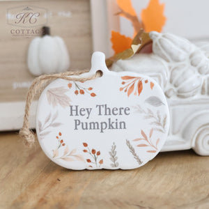 An "Autumn Ceramic Pumpkin Shaped Hanger With Quote," adorned with autumn leaf and berry designs and the text "Hey There Pumpkin," sits on a wooden surface near a white ceramic pumpkin, accompanied by an autumn-themed decor piece in the background.