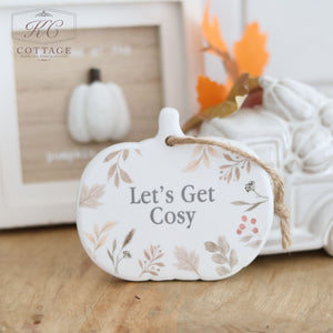 An Autumn Ceramic Pumpkin Shaped Hanger With Quote, adorned with charming floral and leaf designs and featuring the inscription "Let's Get Cosy" in the center, rests on a wooden surface. In the background, additional seasonal decorations include a white ceramic pumpkin and a small white truck carrying fall leaves.