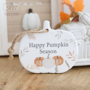 The Autumn Ceramic Pumpkin Shaped Hanger With Quote displays the text "Happy Pumpkin Season" on its white, pumpkin-shaped ceramic surface. This enchanting seasonal decor piece features illustrations of pumpkins and autumn leaves, complemented by a twine loop at the top. It rests on a wooden surface against a backdrop of fall-themed decorations.