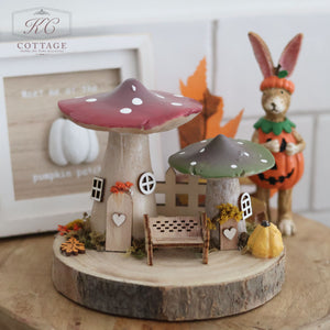 An enchanting autumn-themed decoration, the "Autumn Toadstool Two Cottages Medium," featuring two mushroom houses with red and green caps surrounded by small pumpkins and foliage. A small bench sits between them, while a bunny figurine in a pumpkin costume stands behind. In the background, a sign with the KC Cottage logo evokes the charm of Two Cottages.