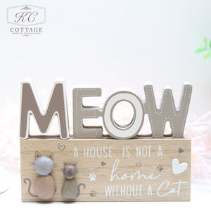 Introducing the Cat Pebble Signs: a charming wooden decor piece featuring large letters that spell "MEOW" and smaller text declaring, "A house is not a home without a cat." Ideal for any cat enthusiast's abode, this sign includes two adorable cat figurines flanking the text. The background showcases a light, pastel design with subtle touches of pink and green. Proudly adorned with the "KC Cottage" logo in the top left corner.