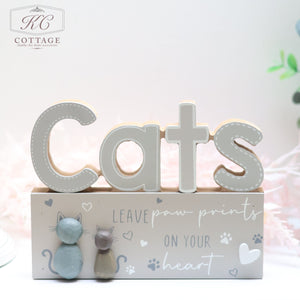 Introducing the Cat Pebble Signs, a decorative wooden sign perfect for any cat lover's home. It features the word "Cats" in large letters and includes two charming cat figurines—one gray and one beige—sitting on a rectangular base. The base is embellished with paw prints, hearts, and the heartwarming phrase "Leave paw prints on your heart" in a beautiful decorative font.