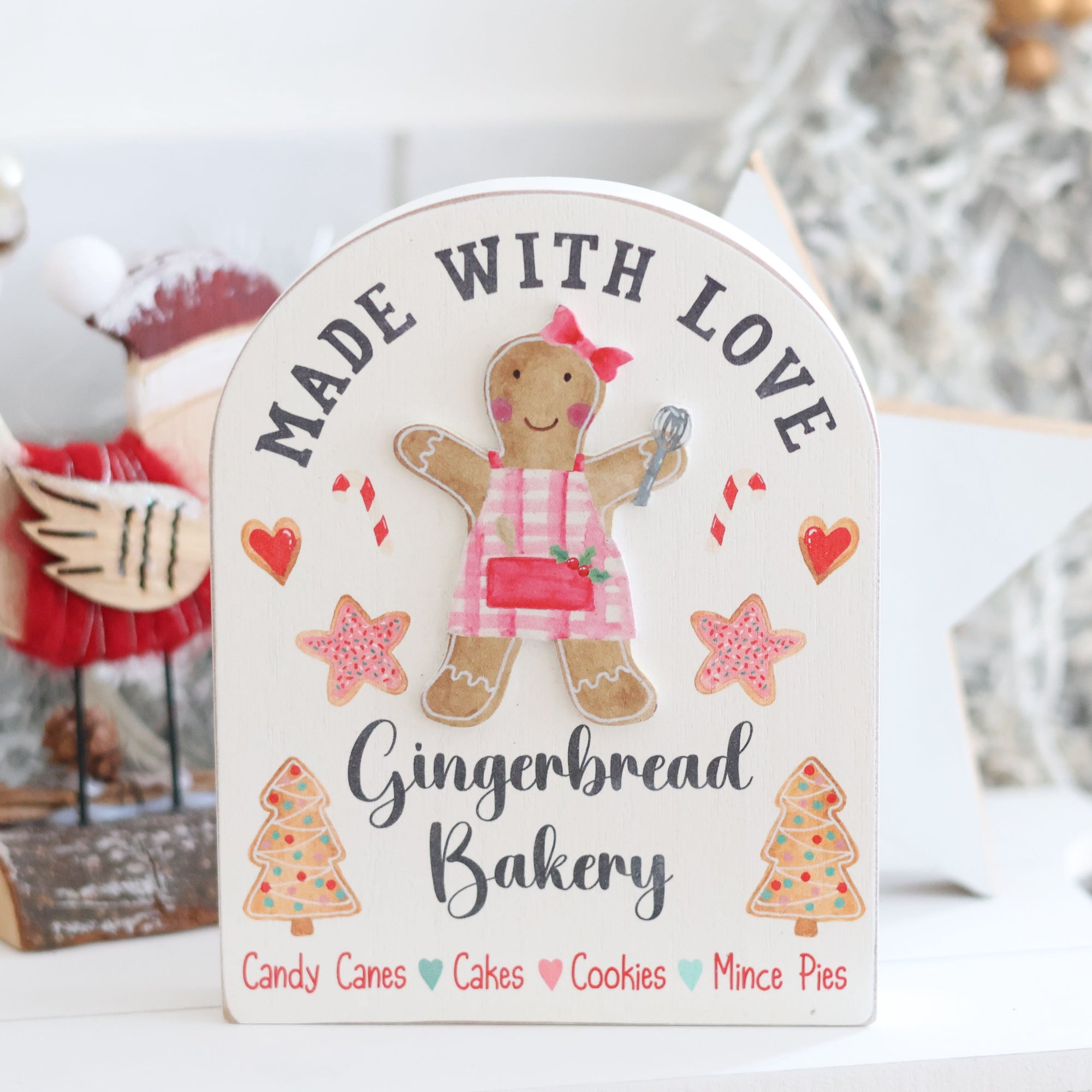 The Christmas Gingerbread Bakery Arched Block Sign showcases a festive gingerbread girl holding a whisk, dressed in a red bow and dress, decorated with hearts and Christmas trees. The text reads: "Made with Love - Gingerbread Bakery," featuring the items "Candy Canes, Cakes, Cookies, Mince Pies." Holiday decorations complete the charming display.