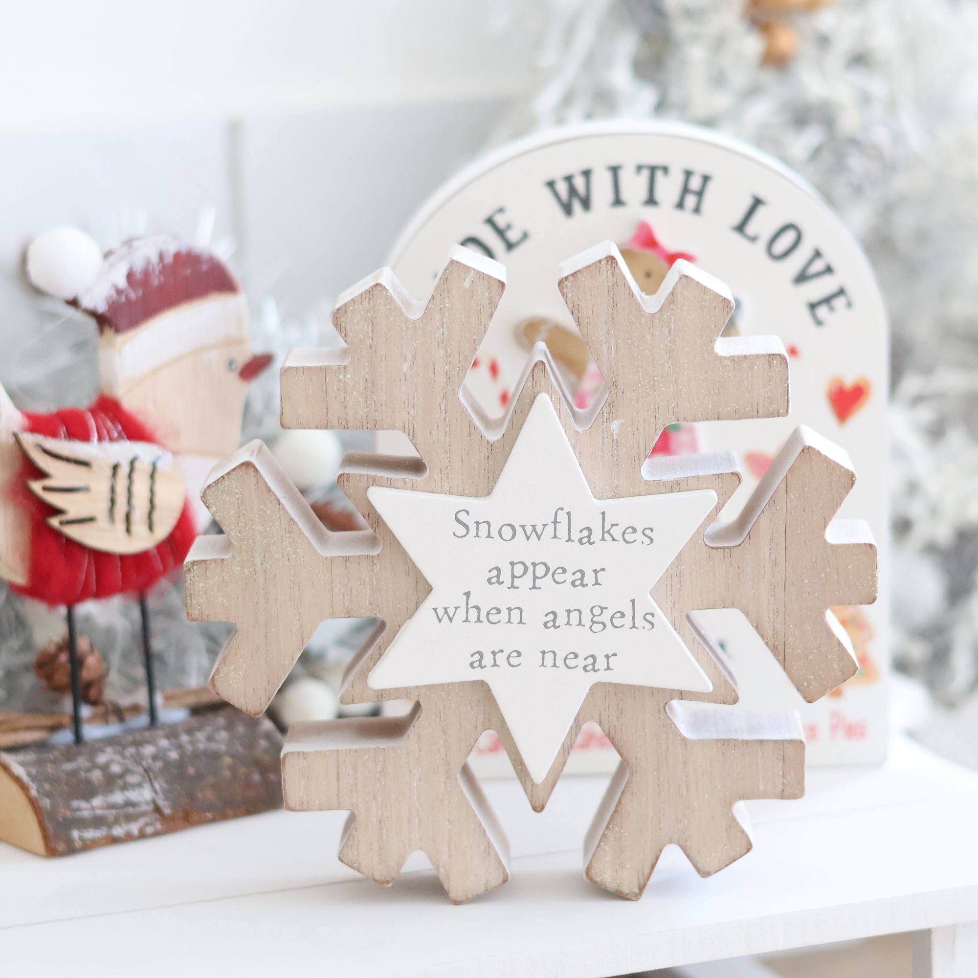 A Christmas Snowflake with Pastel Star decoration featuring the text "Snowflakes appear when angels are near" stands on a white surface. In the background, a festive scene includes a small red and white bird figurine and a "Made with Love" sign, radiating festive cheer.