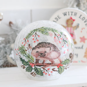 The Christmas Hedgehog LED Ball Crackle Glass showcases a beautifully painted Christmas hedgehog surrounded by sprigs of holly and red berries. The background features blurred festive decorations and a round sign reading, "Made with Love," accompanied by an illustration of a gingerbread figure.