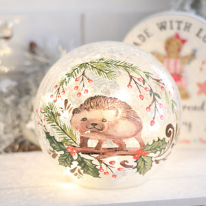 The Christmas Hedgehog LED Ball Crackle Glass showcases a charming painted Christmas hedgehog amid green leaves and red berries. The background features a circular sign with text and an adorable teddy bear illustration, evoking a festive and cozy atmosphere.