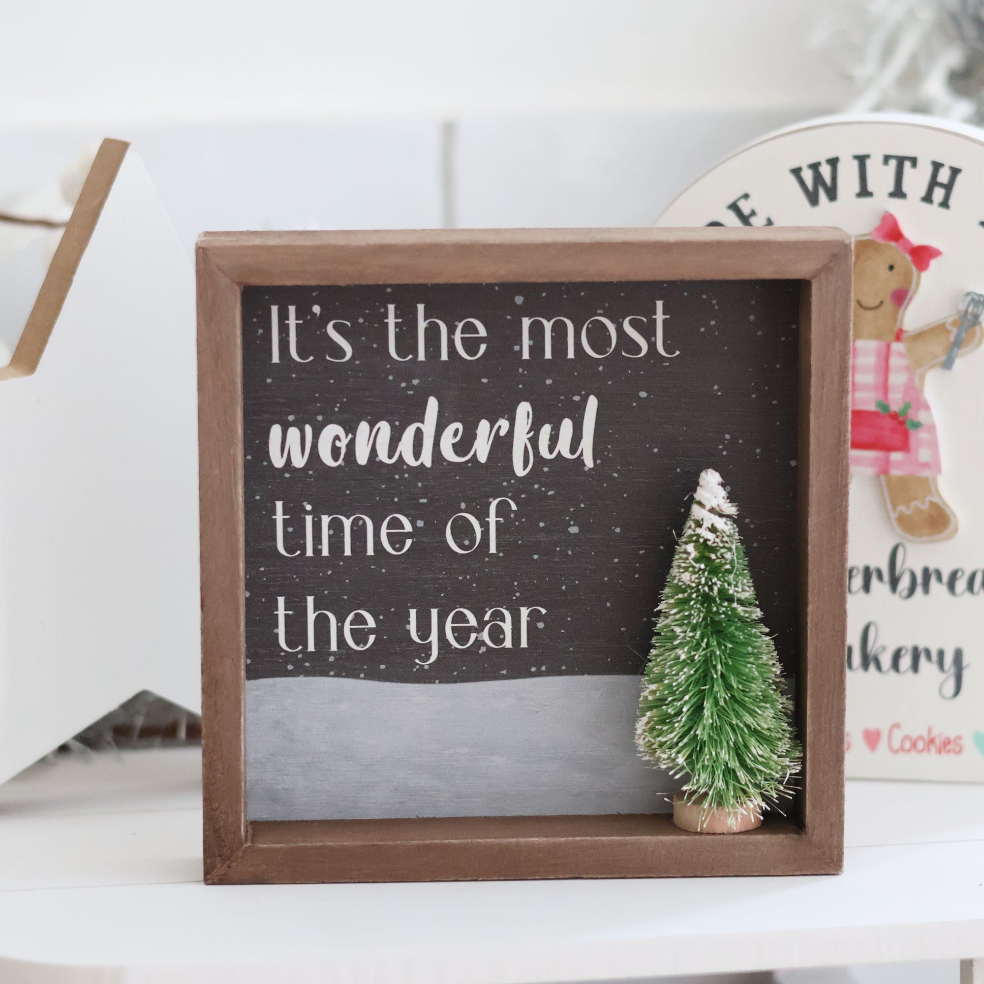 The Wonderful Christmas with 3D Tree Sign showcases the phrase "It's the most wonderful time of the year" in elegant white text against a black background. The sign is beautifully framed and features a small, snow-covered green Christmas tree decoration in front, creating a delightful winter wonderland atmosphere.