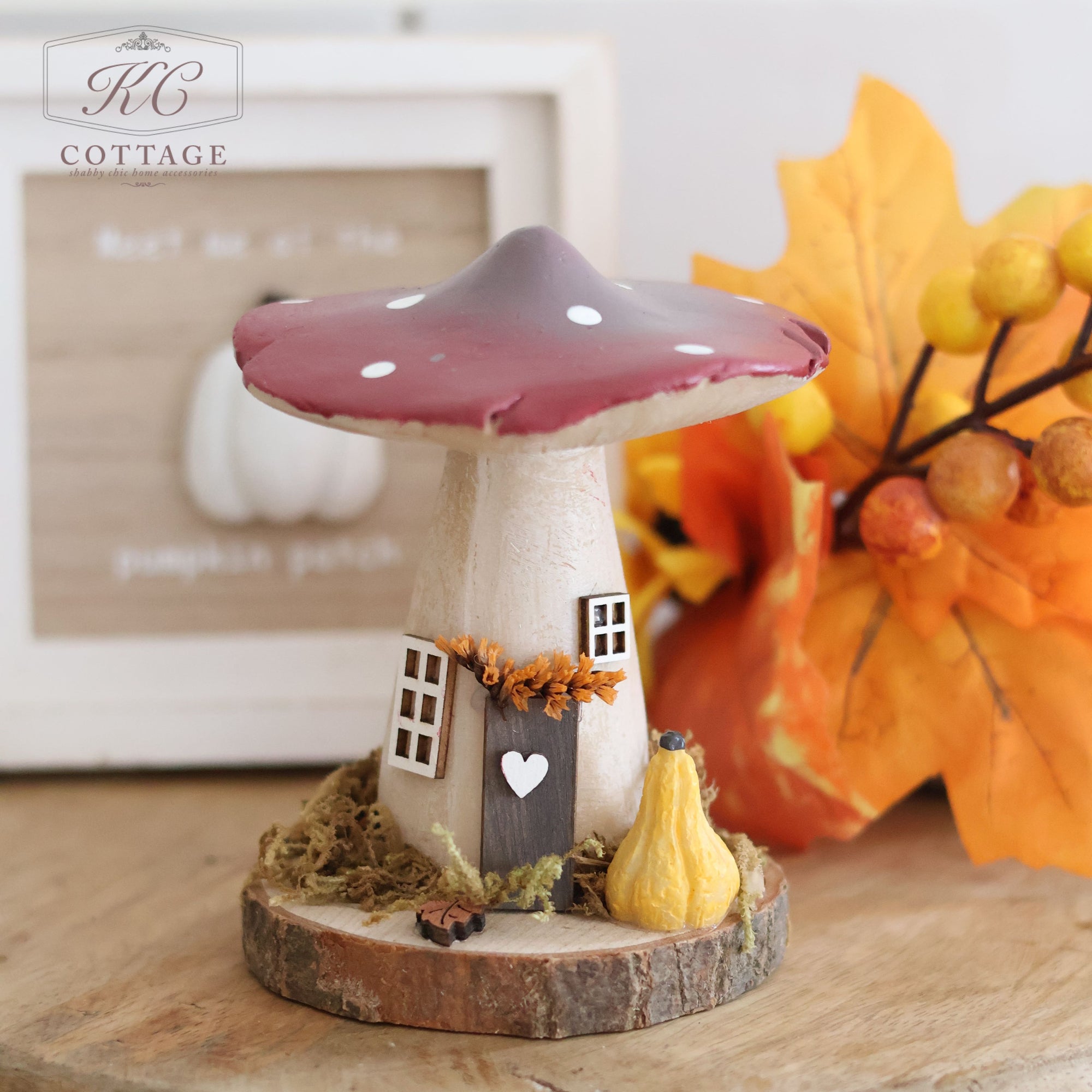 A medium-sized Autumn Toadstool Cottage, featuring a brown-capped mushroom-shaped decoration, sits on a wooden disk base. Its intricate details include small windows, a door with a heart, and adornments of moss and a small pumpkin. An autumn foliage arrangement is partially visible in the background, enhancing its charming appeal.