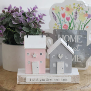 Two small, decorative wooden houses—one pink with a white star and the other gray with a heart—sit side by side, resembling the "I Wish You Lived Next Door Double House Ornament." A sign beneath them reads, "I wish you lived next door," serving as a sweet reminder of friendship. Behind them is a potted plant and another sign that says "HOME SWEET HOME.