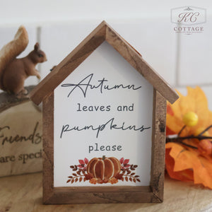 The "Autumn Pumpkin House Framed Sign With Quotes" elegantly showcases the charming phrase "Autumn leaves and pumpkins please" along with a delightful pumpkin graphic at the bottom. Set on a wooden surface beside a squirrel figurine and autumn leaves, this autumn-themed decor piece also reveals part of another framed sign in the background.