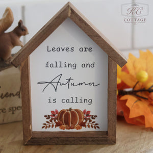 The Autumn Pumpkin House Framed Sign With Quotes reads "Leaves are falling and Autumn is calling" adorned with decorative foliage and an autumn pumpkin illustration underneath. A small squirrel figurine and bright orange leaves enhance the background, creating a cozy centerpiece for any autumn home.