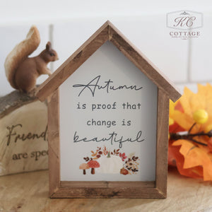 The Autumn Pumpkin House Framed Sign With Quotes sits on a table. Inside the sign, the text reads, "Autumn is proof that change is beautiful," accompanied by illustrations of fall leaves, a pumpkin, and mushrooms. In the background, there is a decorative squirrel and an orange leaf.