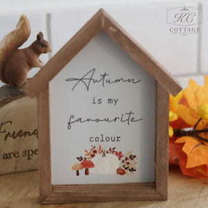 The "Autumn Pumpkin House Framed Sign With Quotes," a wooden sign shaped like a house, displays the text "Autumn is my Favourite Colour" and sits on a table. The bottom of the framed sign is adorned with an autumn-themed illustration featuring pumpkins, mushrooms, and leaves. A small squirrel figurine and artificial autumn leaves are arranged nearby.