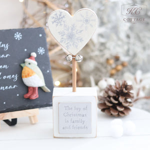 The "Christmas Star & Heart Block With Quote" is a decorative wooden block featuring a heart adorned with snowflake designs and two small bells. An inscription reads, "The joy of Christmas is family and friends." The background includes a pine cone, a folk art bird, and festive holiday decor to enhance the charm of this delightful Christmas decoration.