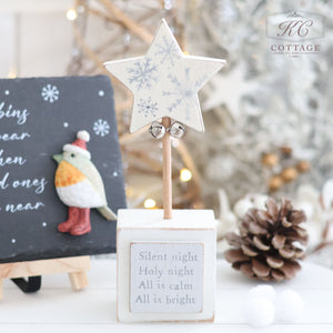 A festive holiday display features the Christmas Star & Heart Block With Quote, which showcases a star with intricate snowflake patterns on a wooden stand, adorned with two small bells. Below it is a heartfelt message that reads, "Silent night, Holy night, All is calm, All is bright." In the background, charming bird decorations and other Christmas ornaments add an extra touch of holiday spirit.