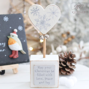 The Christmas Star & Heart Block With Quote is a cozy Christmas-themed decoration featuring a heart atop a wooden block inscribed with "May your Christmas be filled with love, Peace, and Joy." This charming piece includes a small bird ornament and festive backdrop, adding warmth and elegance to the holiday decor while creating an atmosphere filled with inspirational messages.