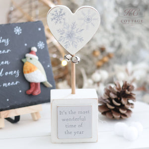 The "Christmas Star & Heart Block With Quote" features a heart-shaped ornament adorned with snowflake designs and small bells, situated on a white wooden block that reads "It's the most wonderful time of the year." The scene is completed with a festive bird figurine, a pinecone, and a chalkboard displaying inspirational quotes and holiday decor in the background.