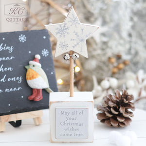 A festive scene featuring the Christmas Star & Heart Block With Quote, decorated with a star on a stand, two small bells, and intricate snowflake patterns. The base bears the heartfelt message, "May all of your Christmas wishes come true." In this charming holiday setup, a pinecone and a chalkboard showcasing a vibrant bird design embellish the background.