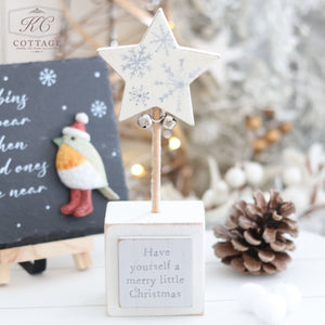 A holiday decor item from the Christmas Star & Heart Block With Quote collection features a wooden star atop a stick in a small block with the text "Have yourself a merry little Christmas." The background includes a chalkboard with a bird design, pinecones, white pom-poms, festive lighting, and other Christmas decorations to inspire joy.