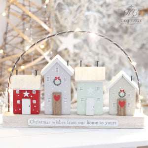 The Christmas Family Wishes Block Wooden Houses With LED Wire Arch features four small block wooden houses in light gray, green, and red, adorned with Christmas wreaths and stars. These charming houses are arranged under a twinkling LED wire arch, and the base reads "Christmas family wishes from our home to yours.