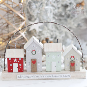 The Christmas Family Wishes Block Wooden Houses With LED Wire Arch is a festive decoration featuring four whimsical block wooden houses, each painted in vibrant Christmas designs, sitting on a wooden base. An LED wire arch frames the houses, while the base displays a sign that reads, "Christmas family wishes from our home to yours.