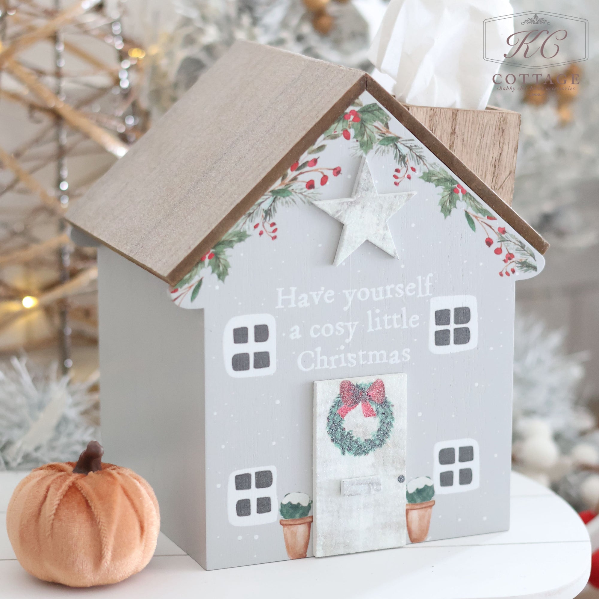 The Christmas Cosy House Tissue Box Holder is an adorable decoration shaped like a small house, adorned with festive Christmas details. It features a wreath on the door and the words "Have yourself a cosy little Christmas" on the front. A small pumpkin sits next to it, set against a background of blurred holiday lights for a charming festive touch.