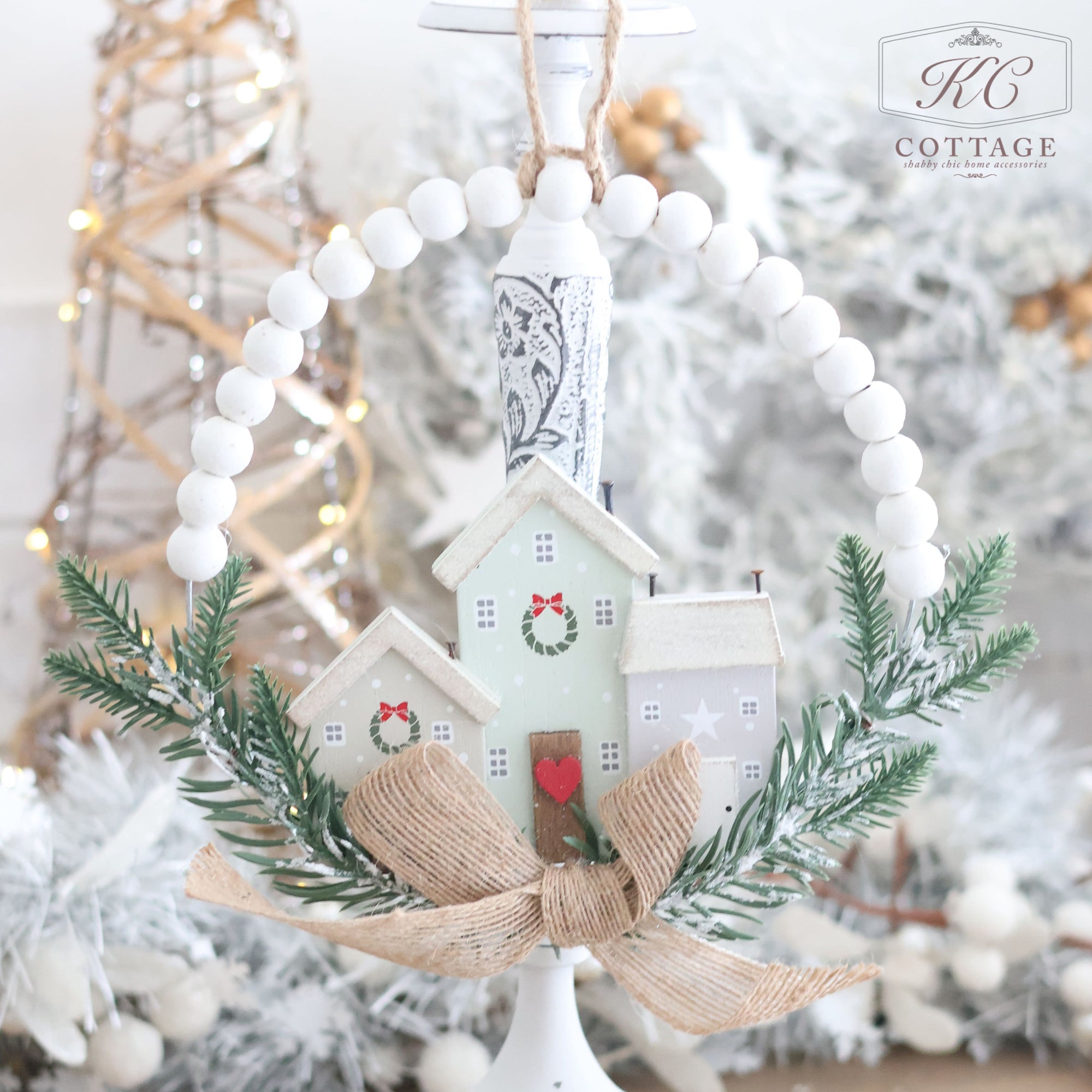 Introducing the Christmas Hanging Houses with Bead Circle Foliage & Bow, a festive ornament showcasing a charming miniature house decorated with a wreath. This delightful piece is encircled by pine branches, accented with a burlap bow and white beads. Set against a backdrop of snowy decorations and warm lights, it's an enchanting addition to your holiday decor.