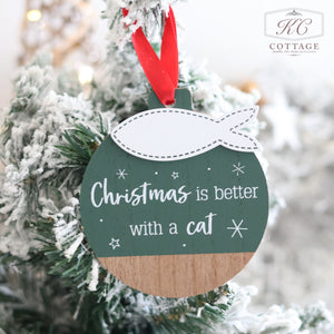 Adorning a snowy tree branch, the Christmas Cat Hanger Green Wood With 3D Fish showcases the phrase "Christmas is better with a cat." This green and wood-textured round ornament features a white cat shape on top and includes a delightful 3D fish detail. It's tied with a red ribbon, and the background highlights the "KC Cottage" logo in the top right corner, perfect for adding festive feline decor to your holiday scene.