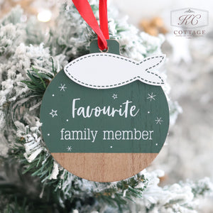 A Christmas Cat Hanger Green Wood With 3D Fish decorating a snow-covered tree. The ornament is circular with a two-tone wooden design—green on top and natural wood at the bottom—accentuated with small white stars. It features a red ribbon and the words "Favourite family member" in white text, making it perfect as festive feline decor.