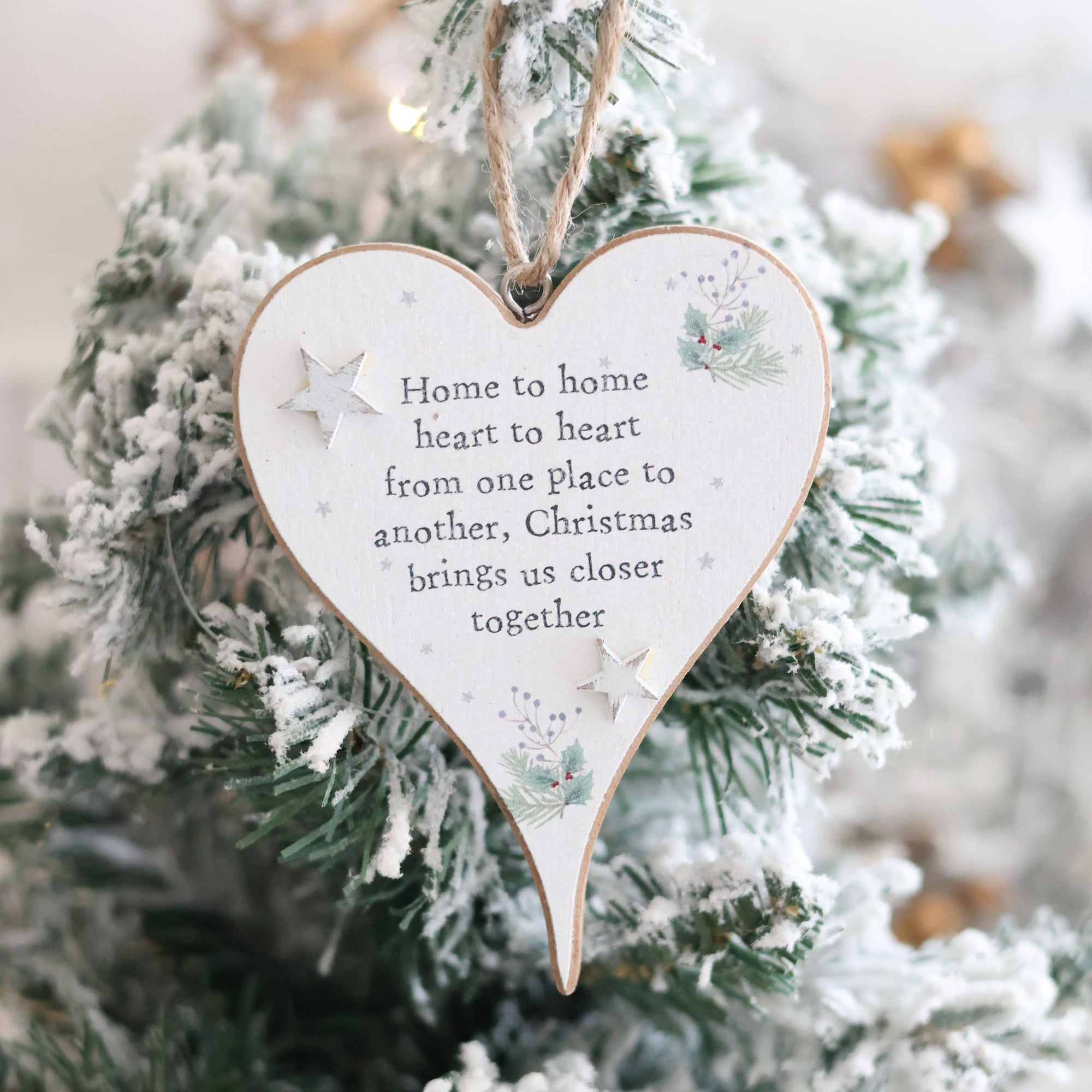 The Christmas Sentiment Heart Hanger With 3D Star, featuring a heartfelt message and adorned with 3D star decorations and festive illustrations, hangs beautifully on a snowy evergreen tree. It reads: "Home to home heart to heart from one place to another, Christmas brings us closer together," bringing festive joy to the holiday season.