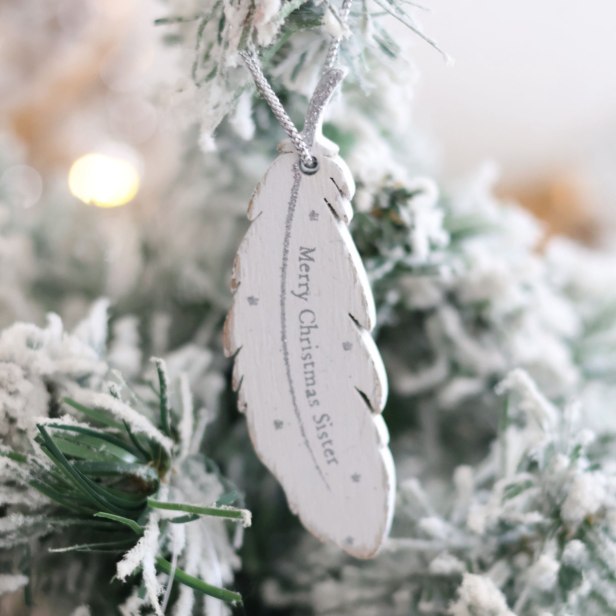 A Christmas Family Sentimental Feather Hanger, featuring a white, feather-shaped design with "Merry Christmas Sister" elegantly printed on it, hangs gracefully from a snowy pine branch. Adorned with small stars and set against a festively glowing background, this cherished memory captures the essence of family love and sentimentality.